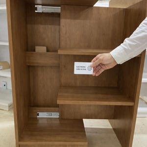 Puede incluir: Un mueble de madera marrón claro con estantes y un cajón. Una mano sostiene una tarjeta con la inscripción "ELECTRONIC CARD ACCESS". El mueble parece ser parte de un conjunto de mobiliario más grande.
