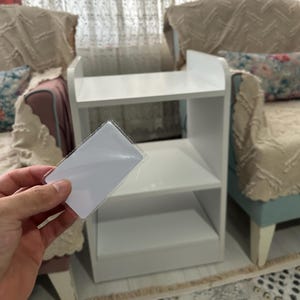 Puede incluir: Una mesa auxiliar blanca de tres niveles con una parte superior y estantes rectangulares. La mano de una persona sostiene una tarjeta rectangular blanca frente a la mesa. La mesa está en una habitación con cortinas estampadas y muebles tapizados.
