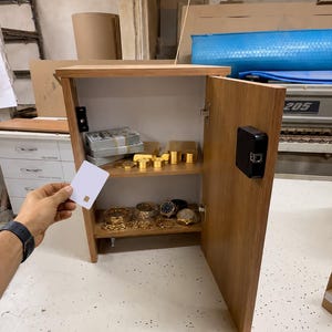 Könnte beinhalten: Ein hellbrauner Wandtresor aus Holz mit elektronischem Kartenleseschloss. Die geöffnete Tür zeigt zwei Regalböden mit Stapeln von Papiergeld, Goldbarren, Goldmünzen sowie verschiedenen goldfarbenen Armbanduhren und Schmuckstücken.