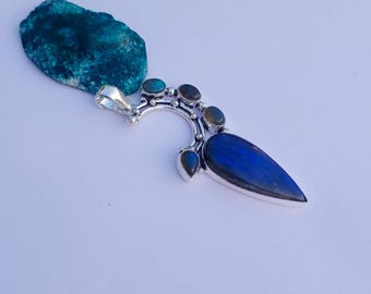 May include: A silver pendant with a large, teardrop-shaped blue stone and smaller, oval-shaped stones in shades of blue and green. The pendant is set against a teal-colored, rough-textured stone.