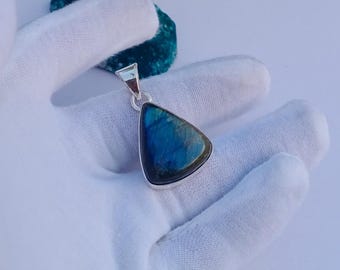 May include: A triangular labradorite pendant with a silver-colored bail. The gemstone displays a vibrant blue and green iridescence. The pendant is held by a gloved hand, suggesting a focus on jewelry or accessories.