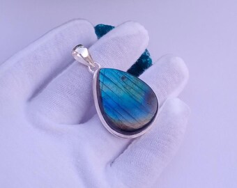 May include: A teardrop-shaped pendant with a vibrant blue and green gemstone. The stone is set in a silver-colored metal frame. The pendant is held by a white-gloved hand, showcasing the gemstone's iridescent quality.