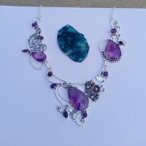 Peut inclure: Un collier en argent orné de trois grosses pierres d'améthyste violettes en forme de goutte et de plusieurs petites améthystes rondes. Le métal présente des motifs en filigrane tourbillonnants et un détail floral près de la pierre centrale.