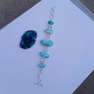 Peut inclure: Un bracelet en argent orné de pierres précieuses ovales et rondes de couleur bleu clair. Le bracelet est doté d'un fermoir à bascule et est présenté sur une surface blanche. Une pierre bleue foncée de forme irrégulière est également présente.