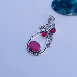 Puede incluir: Colgante de plata con diseño de lágrima, con tres piedras preciosas rojas y un rubí ovalado. El colgante tiene un diseño floral y de remolino decorativo. El colgante está sobre una superficie blanca.