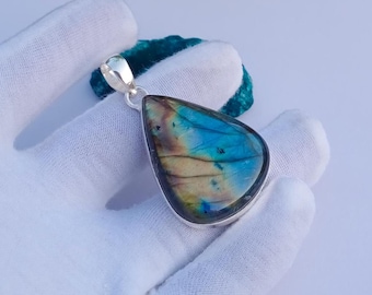 May include: A teardrop-shaped pendant with a silver bail. The pendant features a polished, iridescent stone with blue, green, and gold hues. The stone is set in a silver bezel. The pendant is displayed on a white surface.