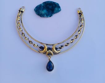 May include: A gold-coloured necklace featuring a crescent moon design and a teardrop-shaped blue gemstone pendant. The necklace has a curved shape with decorative elements, set against a white background.