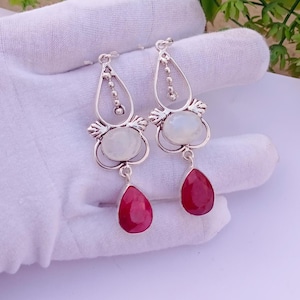 May include: A pair of silver-toned earrings featuring a teardrop design. Each earring has a white, oval-shaped stone and a deep red, teardrop-shaped stone. The earrings are held up by a white glove.