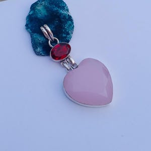 Peut inclure: Un pendentif en argent composé d'une pierre rose pâle en forme de cœur à facettes suspendue sous une pierre rouge foncé de forme ovale. Le pendentif est fixé à une bélière en argent, avec une monture en métal poli retenant les deux pierres.