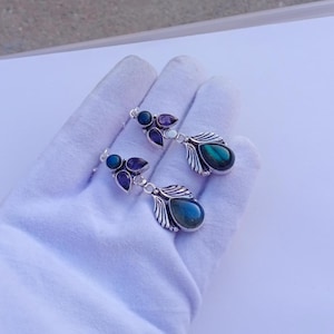 Puede incluir: Un par de pendientes de plata con una piedra azul-verdosa iridiscente en forma de lágrima. Cada pendiente presenta piedras moradas y azules más pequeñas, y un diseño decorativo de hojas. Los pendientes se muestran sobre una superficie blanca.