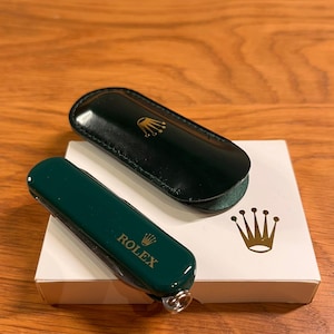 May include: A green Rolex pocket knife and a matching leather case with the Rolex logo. The knife and case are on a white box with the Rolex crown logo. The background is a wooden surface.