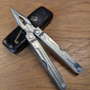 Könnte beinhalten: Ein silbernes Multitool mit Zange, Messer und anderen Werkzeugen, das auf einer Holzoberfläche liegt. Das Werkzeug ist geöffnet und zeigt die Zange. Eine schwarze Ledertasche befindet sich hinter dem Werkzeug.