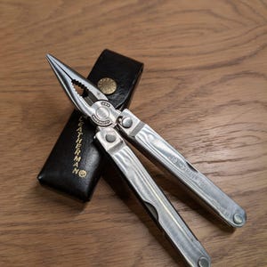 Könnte beinhalten: Ein silbernes Multitool mit Zange, Messer und anderen Werkzeugen, das auf einer Holzoberfläche liegt. Das Multitool ist geöffnet und zeigt seine verschiedenen Komponenten. Ein schwarzes Lederetui mit dem Logo "LEATHERMAN" ist ebenfalls vorhanden.