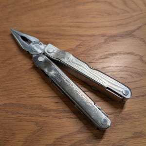 Könnte beinhalten: Ein silbernes Leatherman Super Tool 100 Multitool mit Zange, Messer und anderen Werkzeugen. Das Werkzeug ist geöffnet und zeigt die Zange. Der Text "LEATHERMAN SUPER TOOL 100" ist auf dem Griff zu sehen.