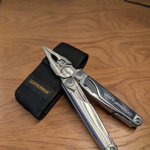 May include: A silver multi-tool with pliers, a knife, and other tools, alongside a black nylon sheath. The multi-tool is branded with "LEATHERMAN SURGE". The sheath is branded with "LEATHERMAN". The tools are on a wooden surface.