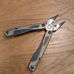 Könnte beinhalten: Ein silbernes und schwarzes Multitool mit geöffneter Zange, das auf einer Holzoberfläche liegt. Der Markenname "GERBER" ist auf dem Griff des Werkzeugs sichtbar. Die Zange ist geöffnet und einsatzbereit. Das Werkzeug ist für verschiedene Aufgaben konzipiert.
