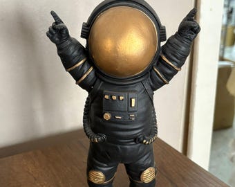 May include: A black astronaut figurine with a gold-colored helmet and accents. The astronaut is standing with arms raised, index fingers pointing upwards. The figurine is on a wooden surface.