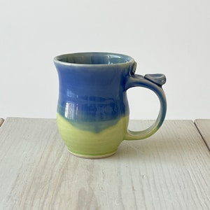 May include: A handmade ceramic mug with a light blue and green glaze. The mug has a unique, slightly curved handle.