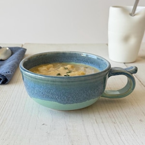 May include: A ceramic soup mug featuring a two-tone glaze with a deep blue upper section and a light turquoise lower section. The mug is filled with a creamy soup garnished with green herbs and includes a sturdy handle on the right side.