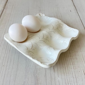 May include: A white ceramic egg holder with a wavy design and embossed leaf pattern, holding two white eggs. The holder has a unique, organic shape, perfect for kitchen decor.
