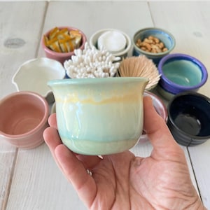 May include: A hand holds a small, light blue ceramic bowl with a yellow rim. The bowl is surrounded by other small, colorful ceramic bowls containing various items, including cotton swabs, nuts, and brushes. The bowls are on a white wooden surface.