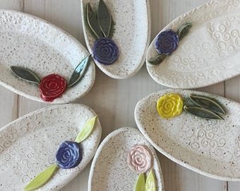 May include: Six oval-shaped ceramic dishes with speckled white surfaces. Each dish is adorned with a colorful ceramic flower and green leaves. The flowers are in red, blue, yellow, and pink. The dishes are arranged on a light wood surface.