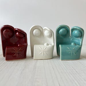 May include: Three ceramic nativity figurines, one in red, one in white, and one in teal. Each figurine depicts a couple standing with a star above a manger.