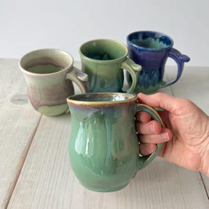 May include: Four handmade ceramic mugs in shades of green, blue, and purple. The front mug is held by a hand and has a glossy green finish with a brown and gold rim. The other mugs have unique color combinations and finishes.