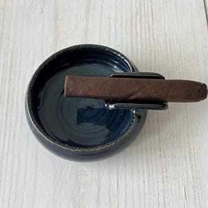 May include: A dark blue ceramic ashtray with a cigar resting on the side. The ashtray has a circular shape with a textured rim. The cigar is brown and appears to be freshly lit.