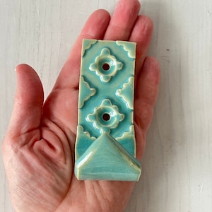 May include: A teal ceramic hook with a decorative pattern and a triangular hook. The hook is held in a hand.