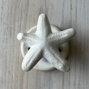 May include: A white ceramic starfish-shaped cabinet knob. The knob has five arms and is mounted on a round, white base with two screw holes. The starfish has a textured surface and is set against a light-colored wooden background.