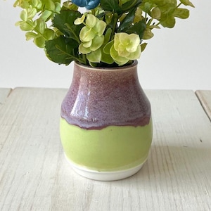 May include: A small ceramic vase with a rounded body featuring a two-tone glaze. The upper portion is a mottled purple and brown, while the lower section is a solid light green. The vase is filled with artificial green foliage and small blue flowers.