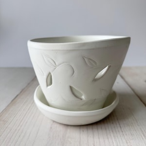 May include: A matte white ceramic planter with a matching saucer. The exterior of the pot features carved leaf motifs and several small, open cutouts shaped like leaves, creating a textured and dimensional appearance on the surface.