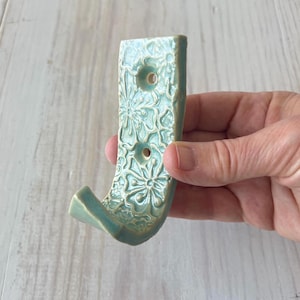 May include: A turquoise ceramic wall hook with a floral pattern. The hook has a J-shape with two holes for mounting. The surface has a textured, embossed design. The hook is held in a hand.