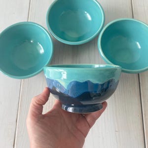 May include: Four ceramic bowls in shades of turquoise and blue. One bowl has a pedestal base and a gradient design from dark blue to light blue. The other three bowls are a solid turquoise color.