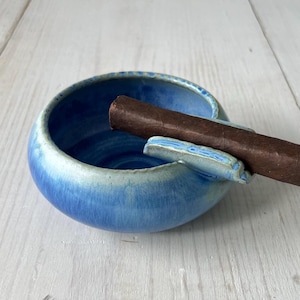 May include: A blue ceramic ashtray with a cigar rest. The ashtray is round with a light blue rim and a darker blue interior. A brown cigar rests in the ashtray. The ashtray is on a light-colored wooden surface.