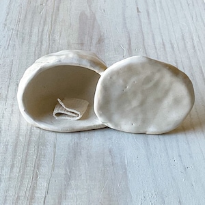 May include: A handmade, off-white ceramic trinket box with a lid, open to reveal a small, folded cloth inside. The box has a rounded, organic shape, suggesting a natural aesthetic. The lid is placed beside the open box.