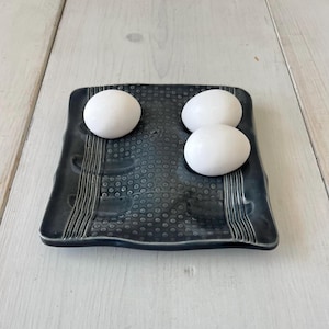 May include: A dark blue ceramic egg holder with a textured surface and three white eggs on top.