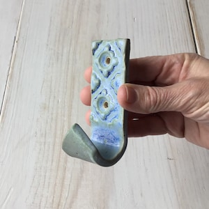 May include: A ceramic hook with a blue and green glaze. The hook has a decorative pattern and two holes for mounting.