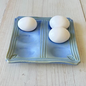 May include: A square ceramic egg tray with a light blue glaze and green trim, featuring four circular indentations. Three smooth white eggs are placed within the tray, with one egg in the top left indentation and two eggs in the right side.