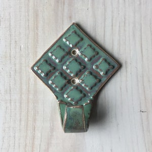 May include: A ceramic hook with a diamond-shaped base and a green glaze. The base has a repeating pattern of squares with rounded edges. The hook is a single, curved piece.