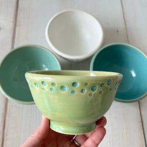 May include: A collection of ceramic bowls in various colors. The foreground bowl is light green with turquoise accents. Other bowls are white, teal, and turquoise. The bowls are on a white wooden surface.