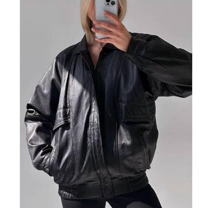 Könnte beinhalten: Schwarze Leder-Bomberjacke mit Kragen, zwei Vordertaschen und geripptem Saum. Die Jacke hat ein klassisches Design mit entspannter Passform, geeignet für verschiedene Anlässe. Das Leder scheint weich und geschmeidig zu sein.