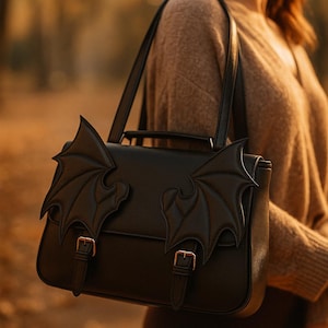 May include: Black satchel bag with bat wing accents. The bag features a flap closure with two buckles and a top handle. The bat wings are attached to the front of the bag. The bag is carried by a shoulder strap.