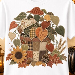 May include: A white short-sleeve t-shirt featuring a central graphic of a patchwork-style basket filled with heart-shaped leaves in autumn colors, a sunflower, a pinecone, an acorn, and wheat stalks, rendered in a quilted fabric texture.
