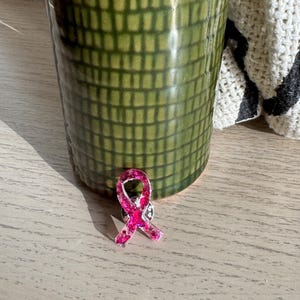 Könnte beinhalten: Ein rosa schleifenförmiger Ansteckpin mit Glitzerakzenten, der auf einer hellen Holzoberfläche liegt. Der Pin ist ein Symbol für Brustkrebs-Aufklärung. Im Hintergrund befindet sich ein grüner, strukturierter Keramikbecher.