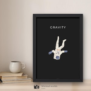 Puede incluir: Lámina artística enmarcada con una ilustración minimalista de un astronauta flotando sobre un fondo negro. La palabra "GRAVITY" está impresa encima del astronauta. El astronauta lleva un traje espacial blanco con guantes azules. Una taza y libros en primer plano.