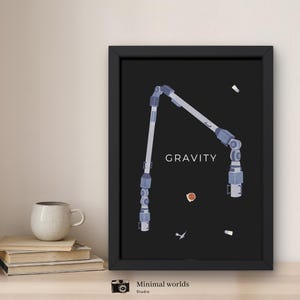 Könnte beinhalten: Schwarz gerahmter Kunstdruck mit einer minimalistischen Illustration eines Weltraumstations-Roboterarms auf schwarzem Hintergrund. Das Wort "GRAVITY" ist zentriert. Der Druck befindet sich neben einem Stapel Bücher und einer weißen Tasse.