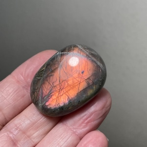 May include: A polished, oval-shaped gemstone with a blend of colors. The stone features a prominent orange and pink center, surrounded by darker gray and purple hues. The surface has a subtle, reflective sheen.