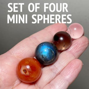 May include: A hand holds a set of four mini spheres. The spheres are in shades of orange, blue, brown, and pink. The text "SET OF FOUR MINI SPHERES" is at the top of the image in white.